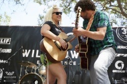 Two musicians perform with acoustic guitars on a small outdoor stage during a festival or community event.