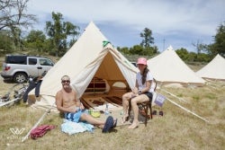 Two people relax outside canvas tents at a campsite during an outdoor cycling or adventure event.