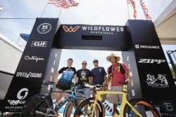 Participants stand with bicycles in front of a sponsor backdrop at an outdoor cycling event.