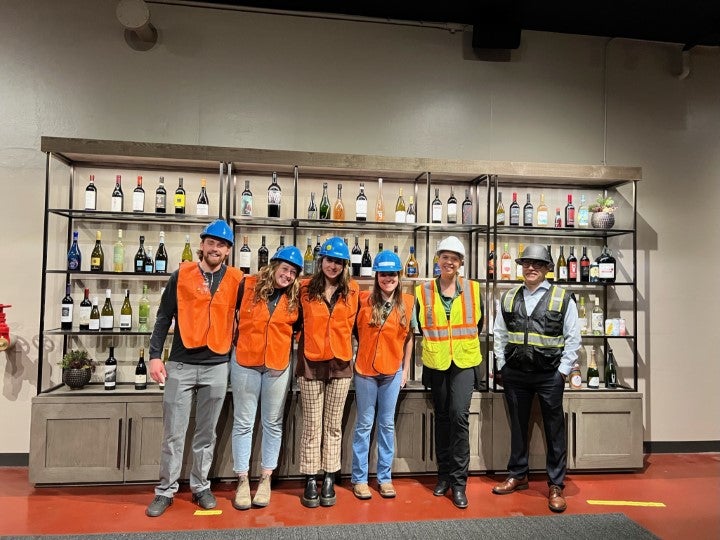 Students wearing hard hats and safety vests during a winery production facility tour in front of wine bottle displays.