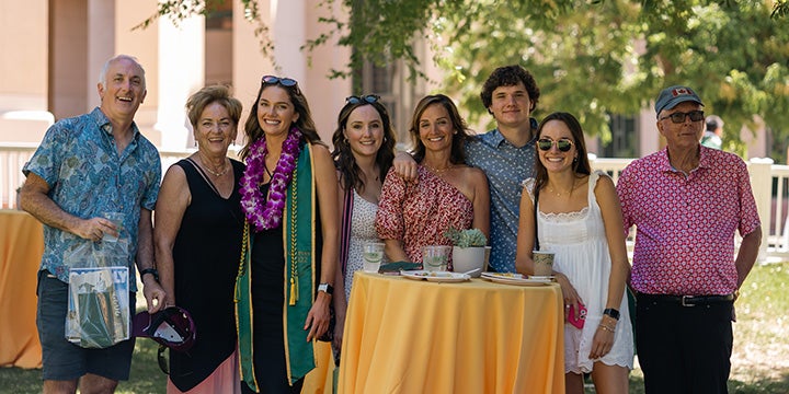 Family and friends gathered around a graduate wearing a green commencement stole and flower lei during an outdoor university graduation celebration.