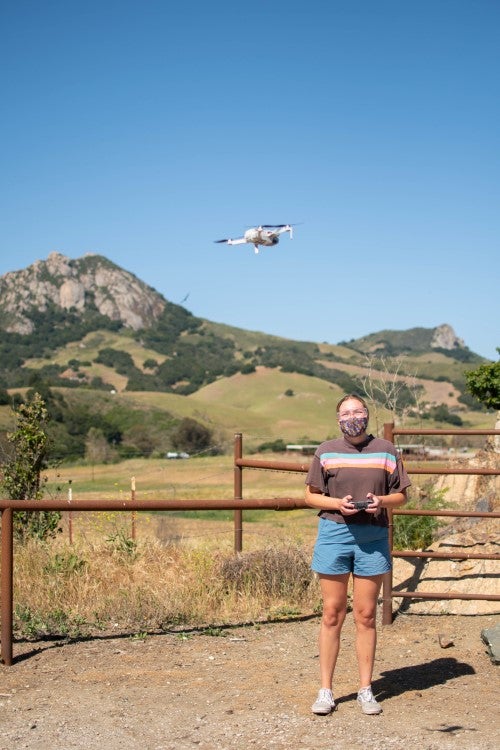A person operates a drone controller outdoors while a drone flies overhead, with hills and open landscape in the background.