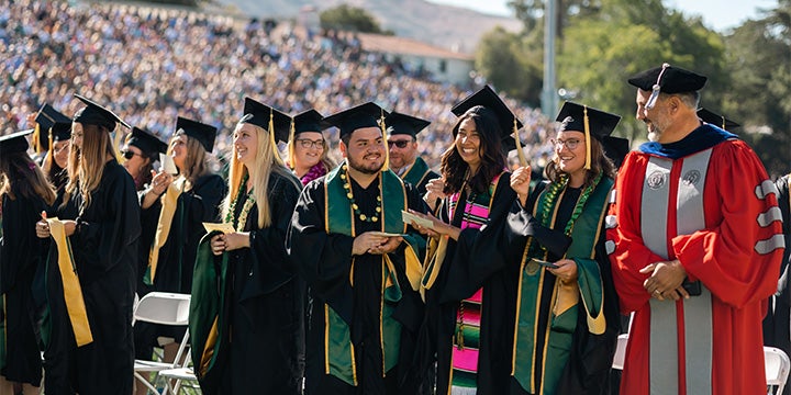 Graduates in caps and gowns standing together during a university commencement ceremony in a crowded stadium alongside faculty members in academic regalia.