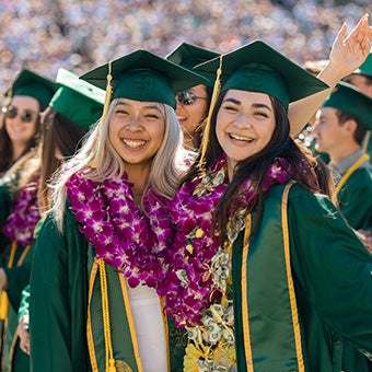 Two smiling graduates wearing green caps and gowns with flower leis posing together during commencement celebrations.