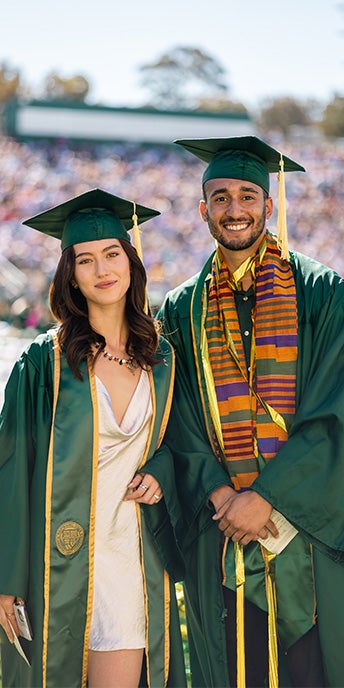 Two university graduates posing together in green commencement regalia, one wearing a colorful cultural stole, at an outdoor graduation ceremony.