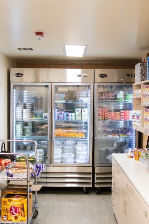 Campus food pantry or food storage space with refrigerated shelves stocked with fresh produce, prepared meals, and packaged food items.