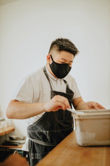 A person wearing a face mask and apron prepares food at a kitchen counter while working with a mixing container.