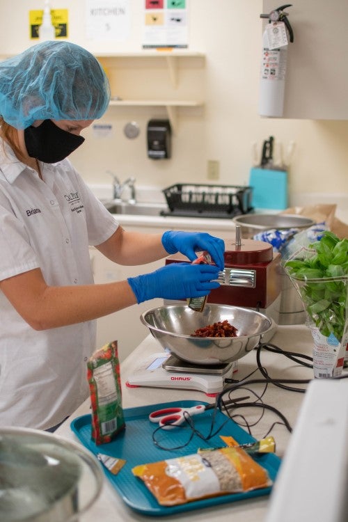 Student in food safety attire preparing ingredients and measuring food components in a commercial kitchen or nutrition science lab.