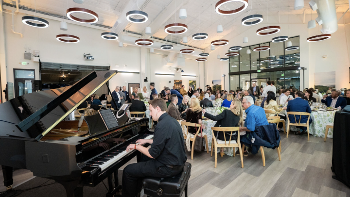 A pianist performs on a grand piano during a large indoor banquet or reception, with guests seated at round tables in a modern event space featuring circular ceiling lights.