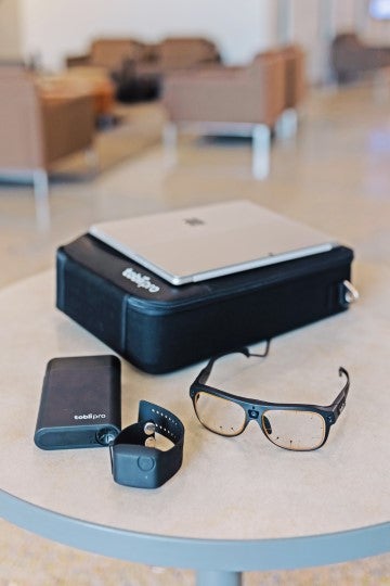 Assistive technology devices, including smart glasses and hearing devices, are displayed on a tabletop in a modern indoor space.