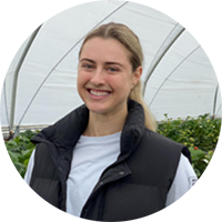 A smiling person wearing a black vest and white shirt stands inside a greenhouse surrounded by rows of plants.