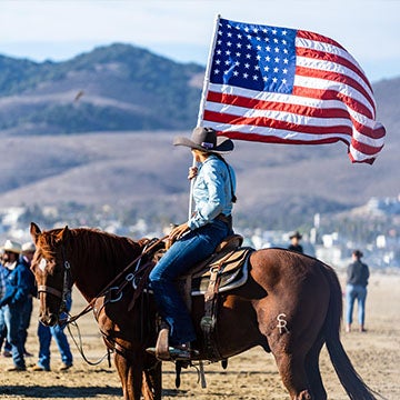 Rider in western attire on horseback carrying a large American flag during a rodeo event, with spectators and rolling hills visible in the background.
