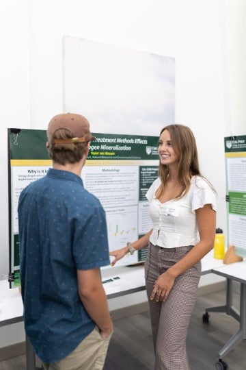 A student discusses a research poster with another attendee during an academic symposium or poster presentation.