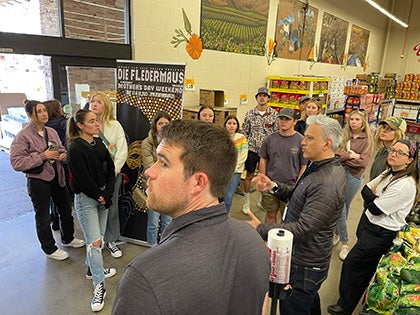 Students gathered inside a grocery or market setting while listening to a speaker during a sustainability, food systems, or agricultural industry educational tour.