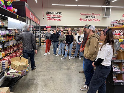 Group of people standing inside a grocery store while listening to a speaker during a guided tour or educational visit.