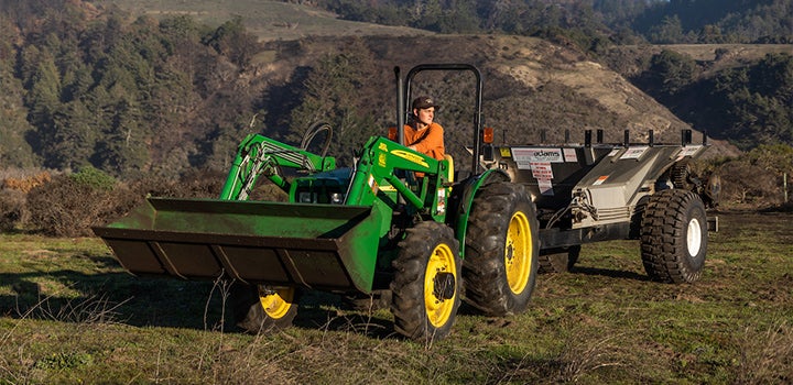 Person driving a green tractor with a front loader attached while towing agricultural equipment across a grassy field with hills and trees in the background.