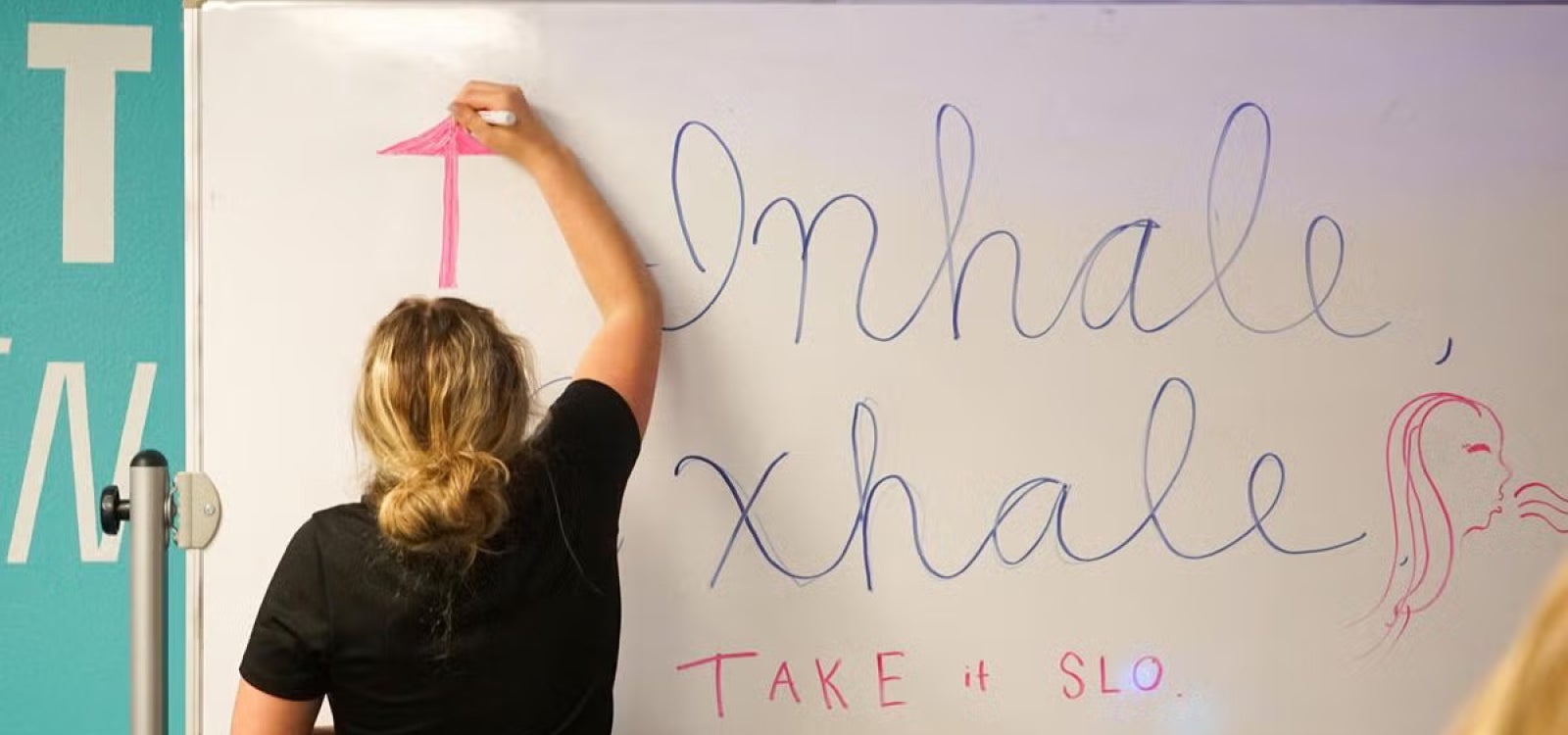 A student draws an arrow point upward on a white board. On the white board are written the words "Inhale" "Exhale" above "Take is SLO".