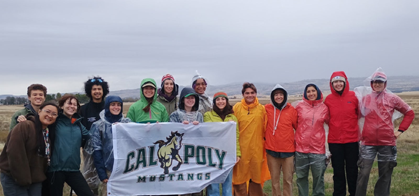 Students wearing parkas and rain jackets hold a Cal Poly Mustangs flag outside in the rain. 