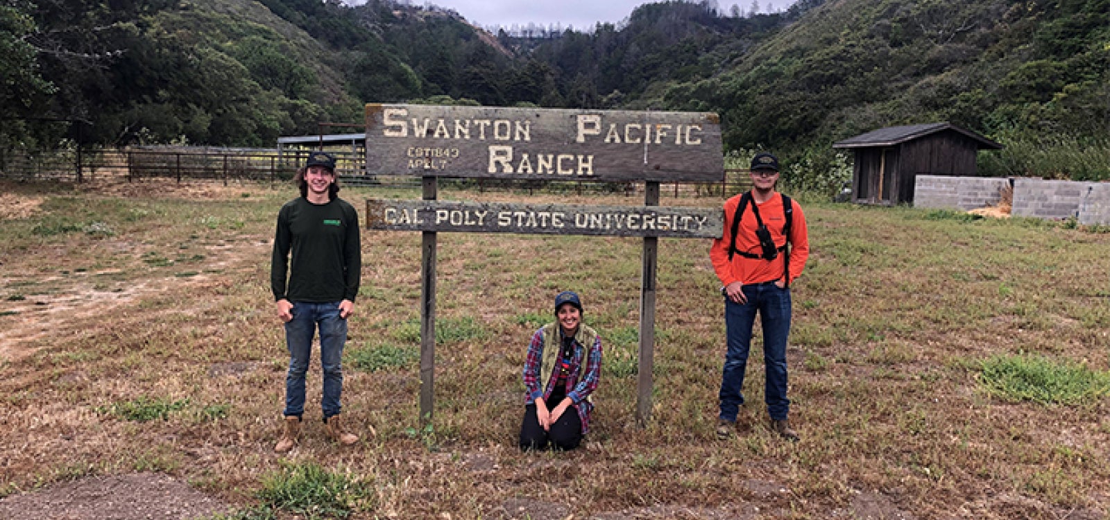 Three students pose for a photo with the Swanton Pacific Ranch sign. Students stand on the left and right of the sign and another squats in front of the sign. 