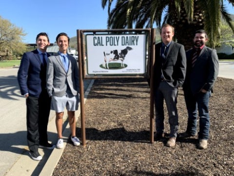 Four men stand beside a Cal Poly Dairy sign on a sunny day, posing for a photo near palm trees and landscaped grounds.