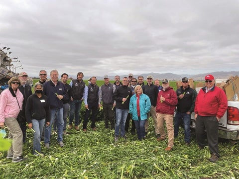A large group of people stand in a harvested field holding produce, posing together during an agricultural tour or field day.