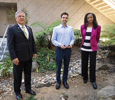Three professionals stand together in a landscaped courtyard with plants and stone groundcover during a formal campus or organizational visit.
