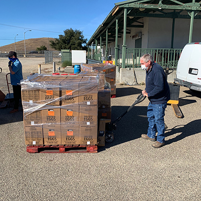 A worker wearing a mask uses a pallet jack to move a large shrink-wrapped pallet of egg cartons outside an agricultural or warehouse facility.