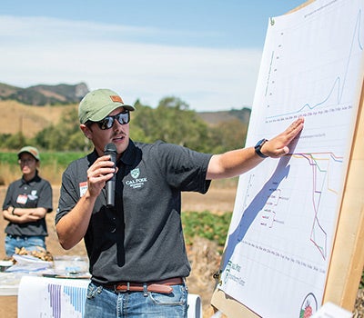 A presenter speaks into a microphone while pointing to a large data chart during an outdoor agricultural field day.