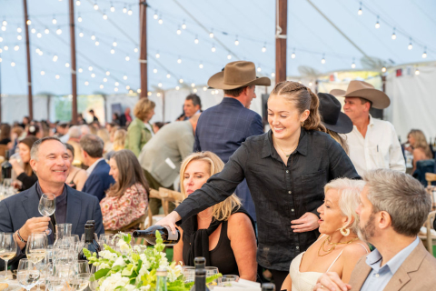 A server pours wine for guests seated at a long dinner table under string lights inside a large event tent.