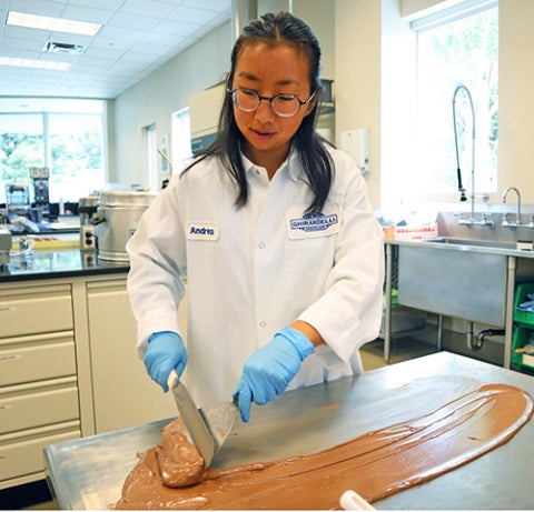 A researcher wearing a lab coat and gloves spreads melted chocolate on a metal surface inside a food science laboratory.