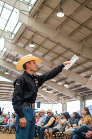 A man in a cowboy hat gestures while holding papers as he addresses a seated audience inside a large livestock or auction pavilion.