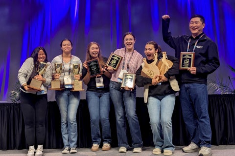Five students and a professor smile as they stand on stage with arms full awards during a Dairy Products Judging competition. 