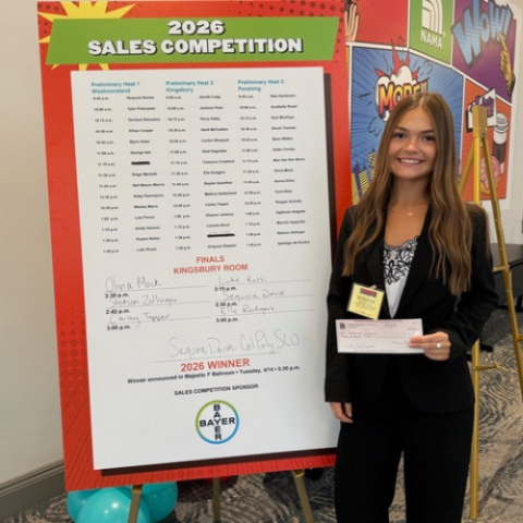 A student stands next to a leaderboard for a national sales competition. 