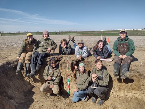 A group of nine students, that make of the Cal Poly Soil Judging Team, pose for a photo while sitting in a large hole in the ground in a field. 