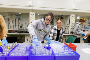 Two students wearing safety goggles work together in a laboratory, handling plastic materials and organizing equipment on a lab bench.