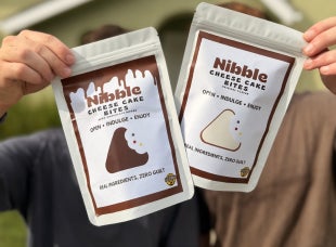 Two hands hold up packages of Nibble Cheese Cake Bites. 