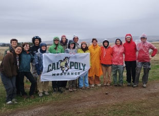 Students wearing parkas and rain jackets hold a Cal Poly Mustangs flag outside in the rain. 