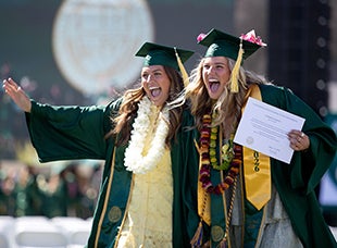 Two female student smile and wave at their family and friends during commencement. 