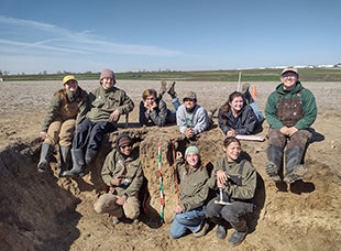 A group of nine students, that make of the Cal Poly Soil Judging Team, pose for a photo while sitting in a large hole in the ground in a field. 