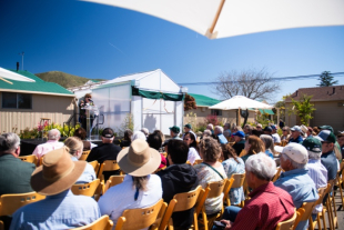 A group of people sitting in chairs while someone speaks on a microphone at the Cal Poly greenhouse
