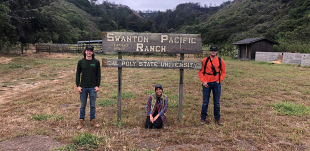 Three students pose for a photo with the Swanton Pacific Ranch sign. Students stand on the left and right of the sign and another squats in front of the sign. 
