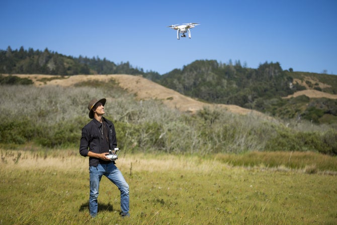 A person standing in a grassy field operates a drone hovering overhead with hills and trees in the background.
