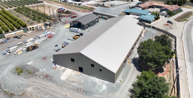 An aerial view of a large industrial building under construction surrounded by equipment, materials, and nearby farmland.