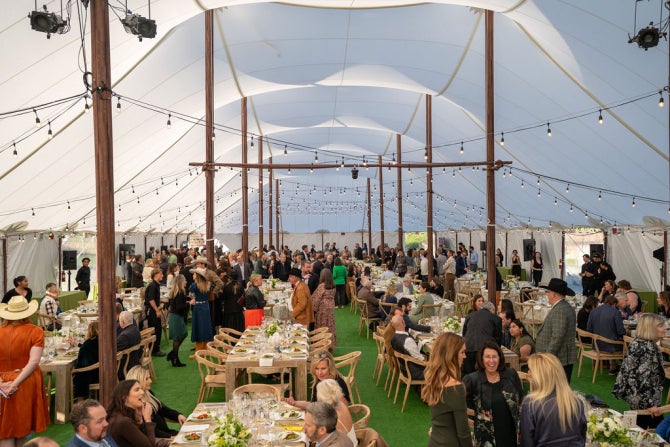 A wide view of guests mingling and dining at decorated tables inside a spacious white tent with hanging string lights and wooden support poles.