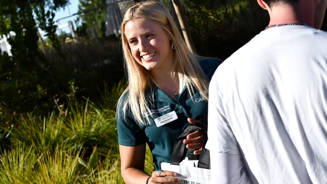 A student in a green Cal Poly polo shirt smiles at passers by. 