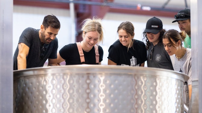 Five students and one professor peer over the top of a large fermentation tank to observe its contents.
