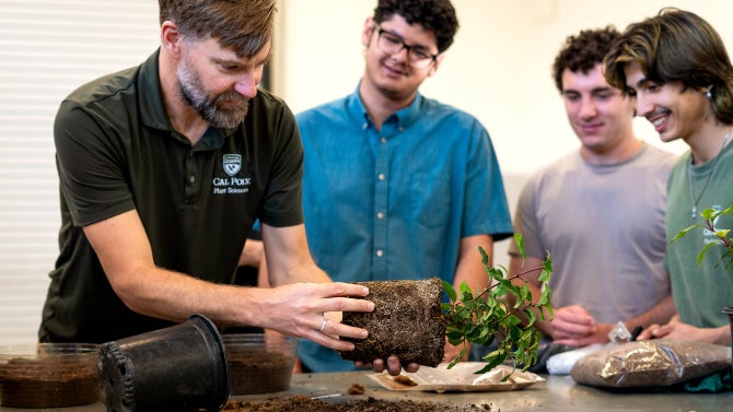 A professor holds a plant, that was just removed from a pot, in front of three students. 