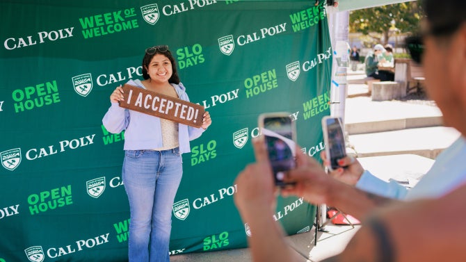 A student gets her picture take as she holds a sign the reads "I Accepted" in front of a Cal Poly Open House step and repeat backdrop. 