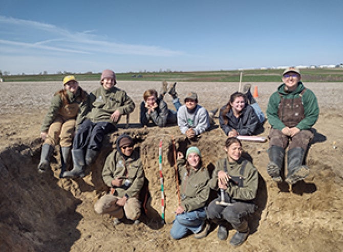 A group of nine students, that make of the Cal Poly Soil Judging Team, pose for a photo while sitting in a large hole in the ground in a field. 