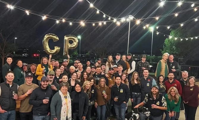A large group of Cal Poly alumni pose for a photo under a dark night sky with hanging lights. 